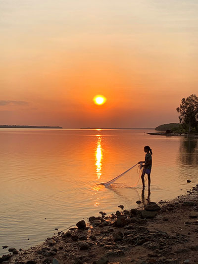 ‘Living in Aurukun’ photography competition. The image is of a beautiful sunset over water and the silhouette of a woman fishing with a net over still water.